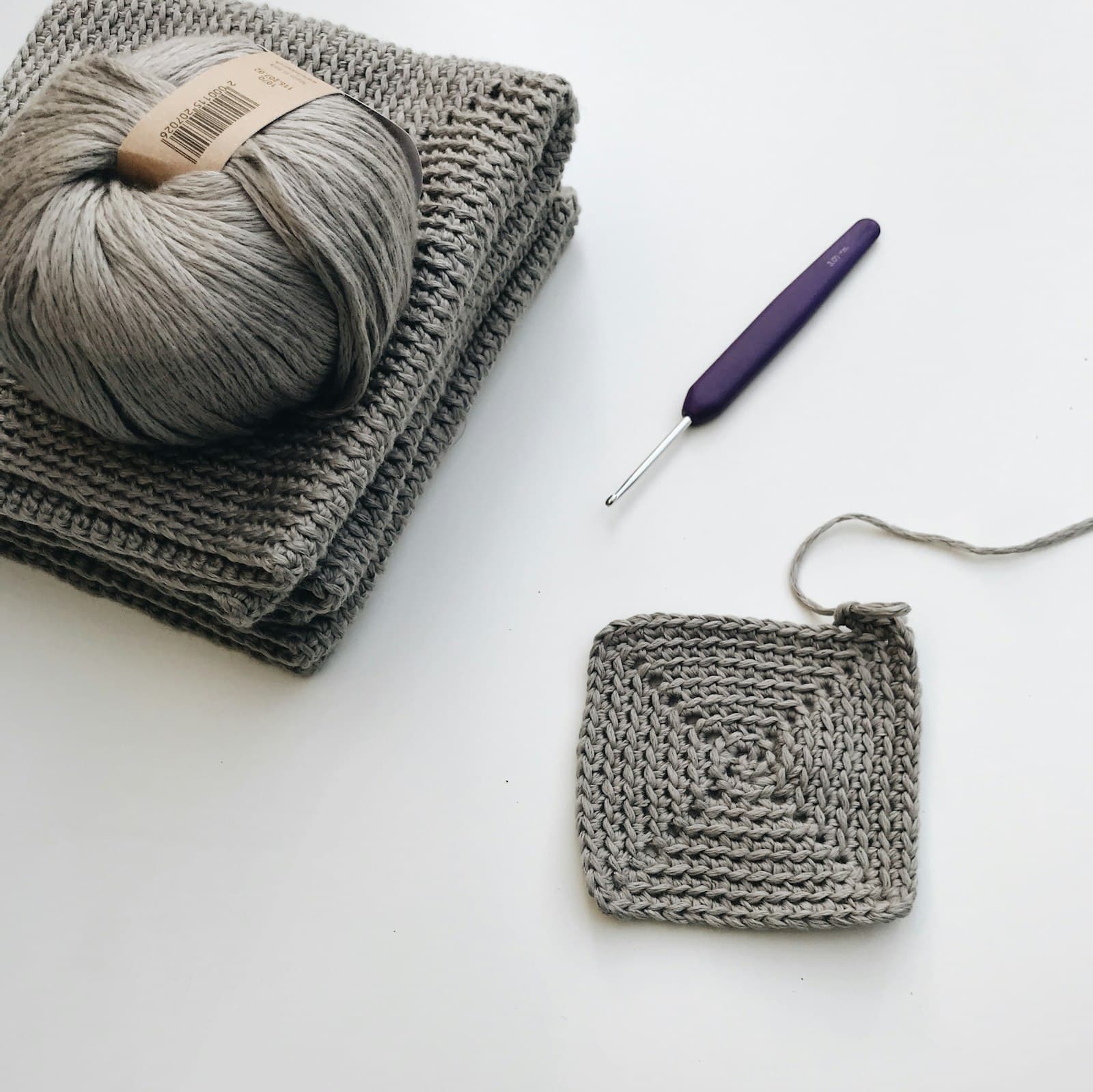 A ball of gray yarn, a purple crochet hook, and a small crocheted swatch laid out on a table