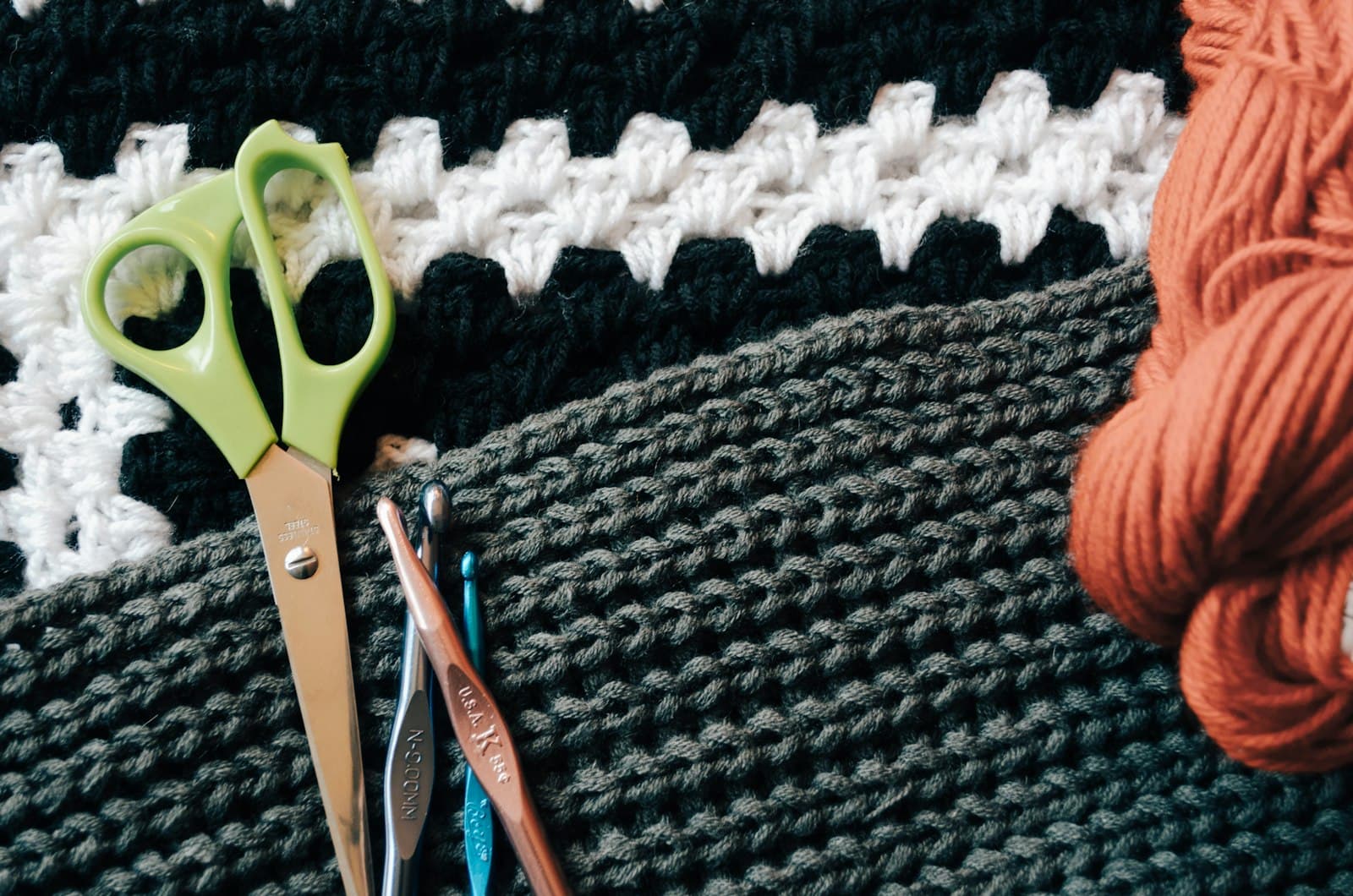 Green-handled scissors and two crochet hooks resting on black, white, and gray crocheted fabric next to a skein of orange yarn