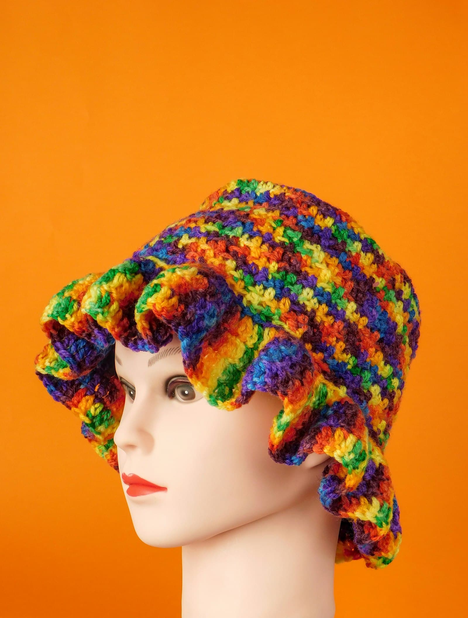 A rainbow-striped crocheted bucket hat with a ruffled brim, worn on a mannequin head against an orange background