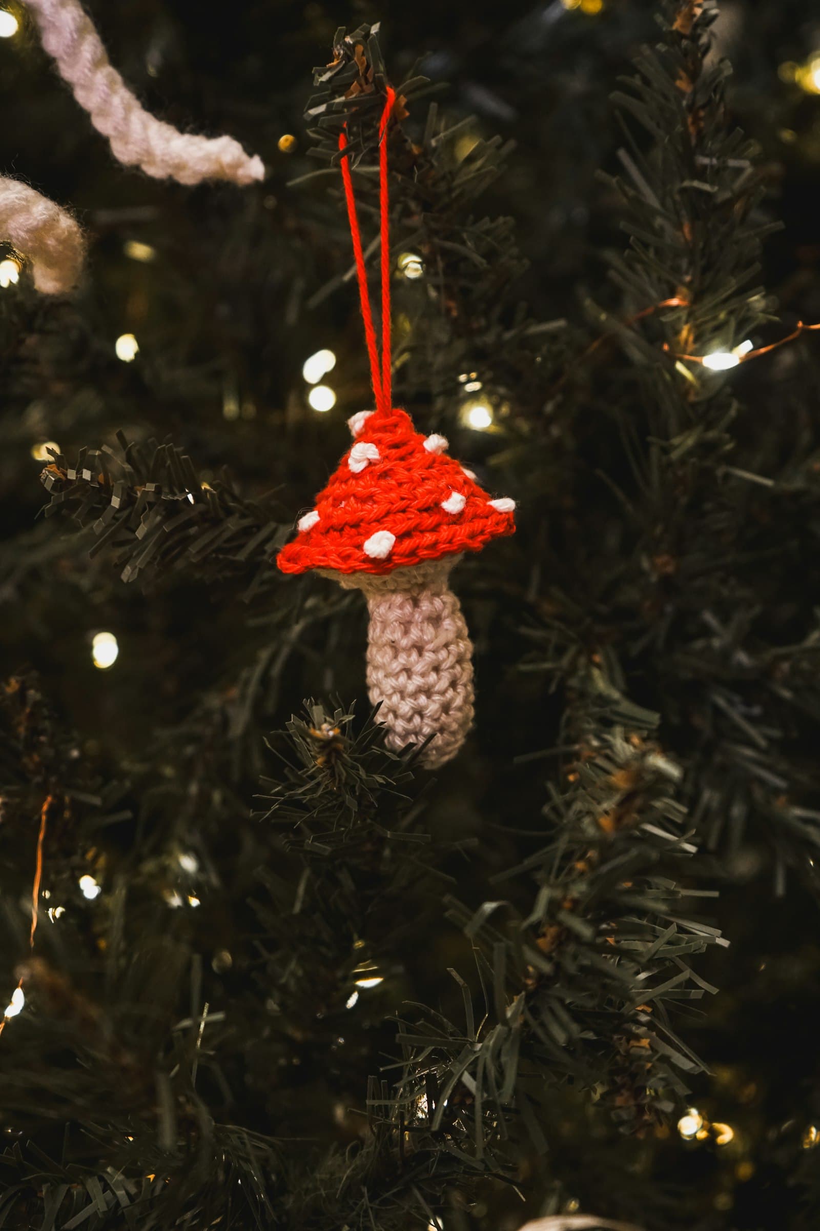 A small red and white crocheted toadstool ornament hanging from a lit Christmas tree branch