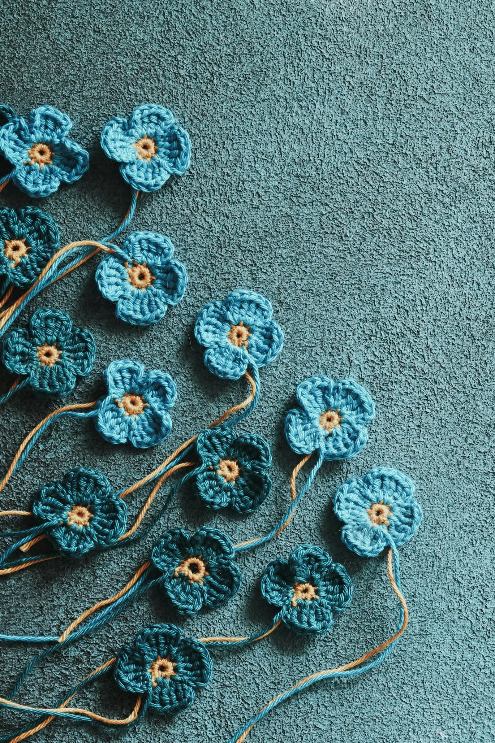 Several small blue crocheted flowers with tan centers, scattered on a teal textured surface