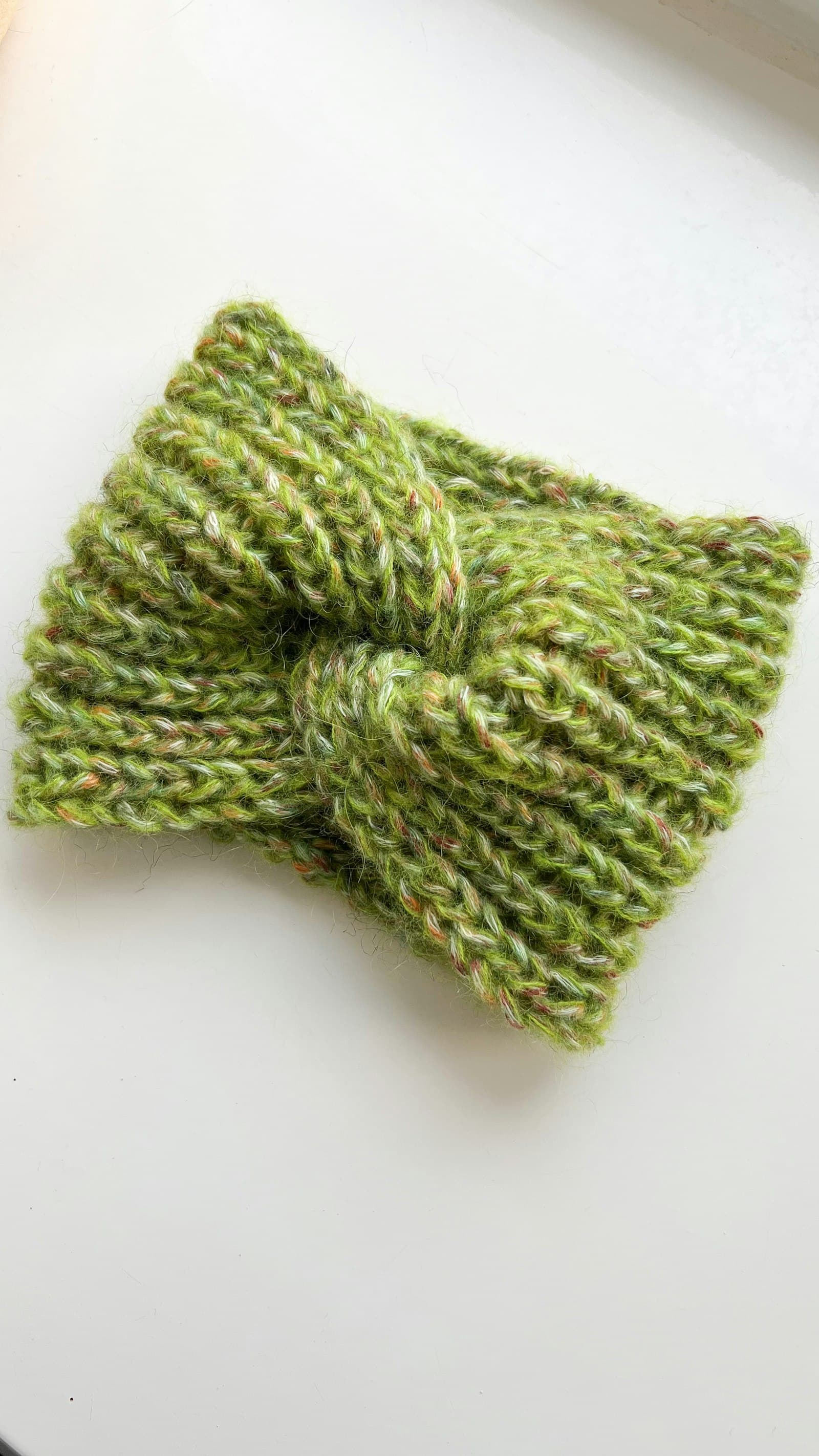 A green knit headband with a twisted knot detail, laid flat on a white surface