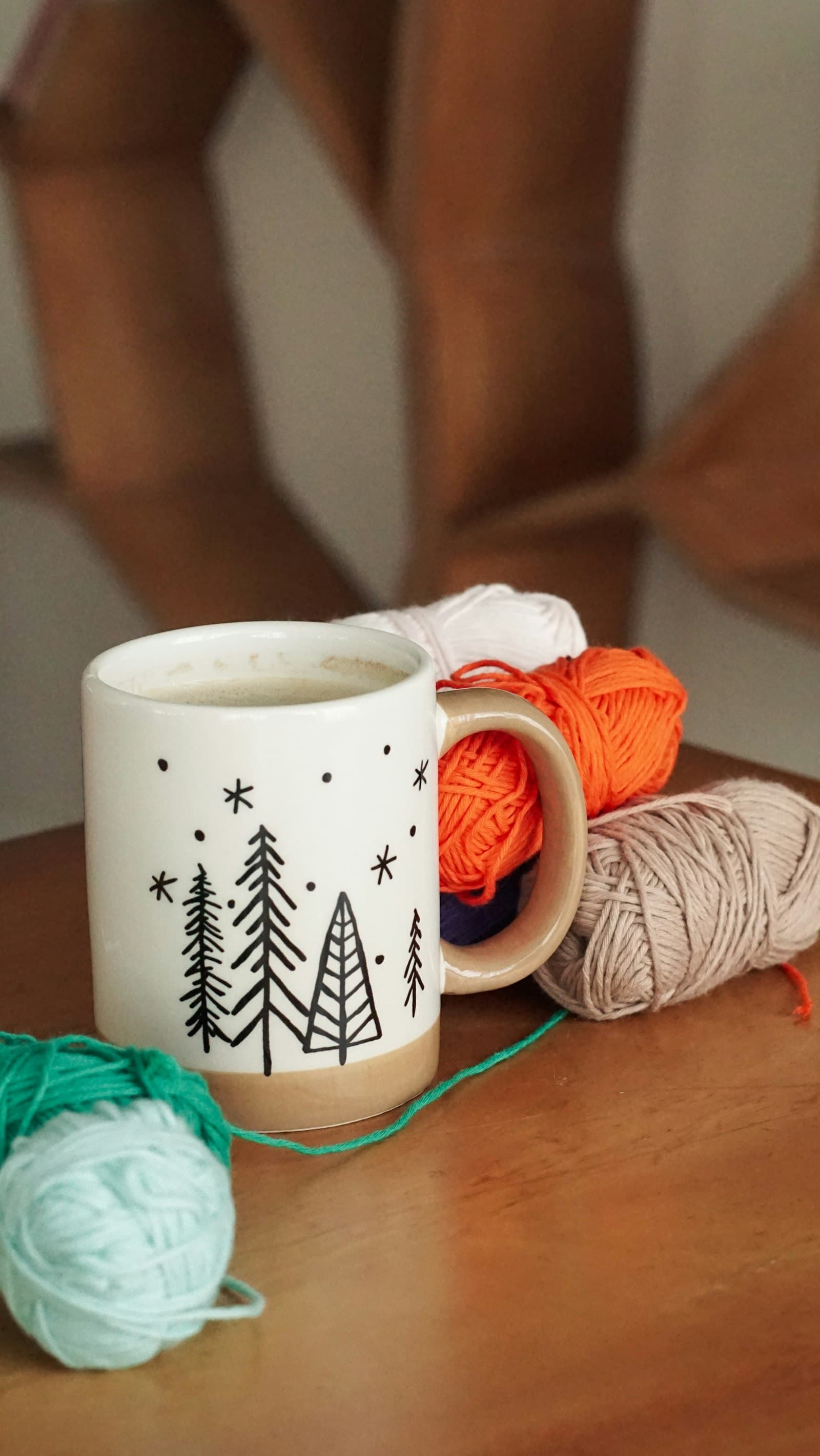 A white mug decorated with pine trees, sitting on a wooden table next to balls of orange, cream, and mint yarn