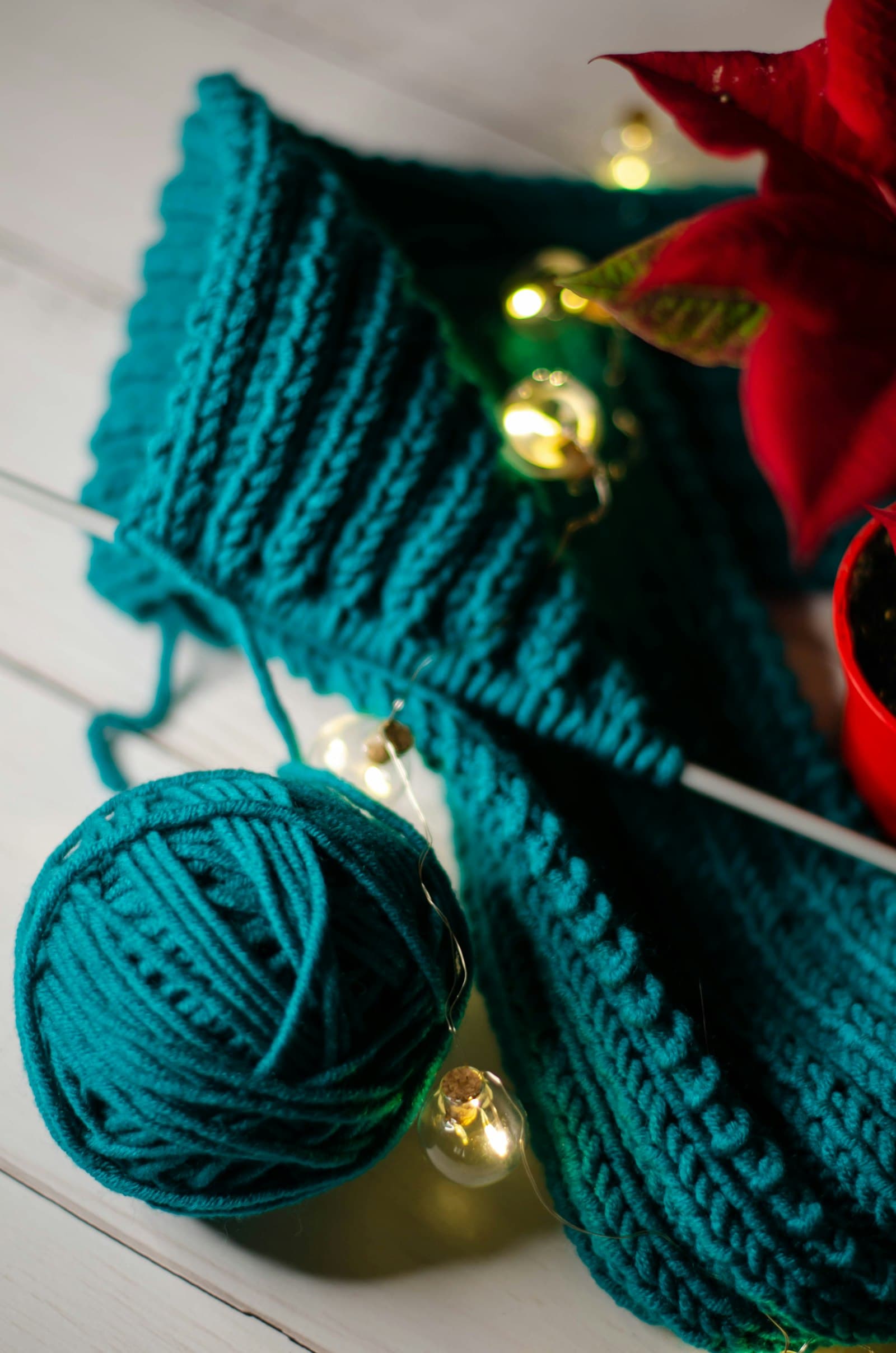 A chunky teal crocheted scarf draped near string lights and a poinsettia plant