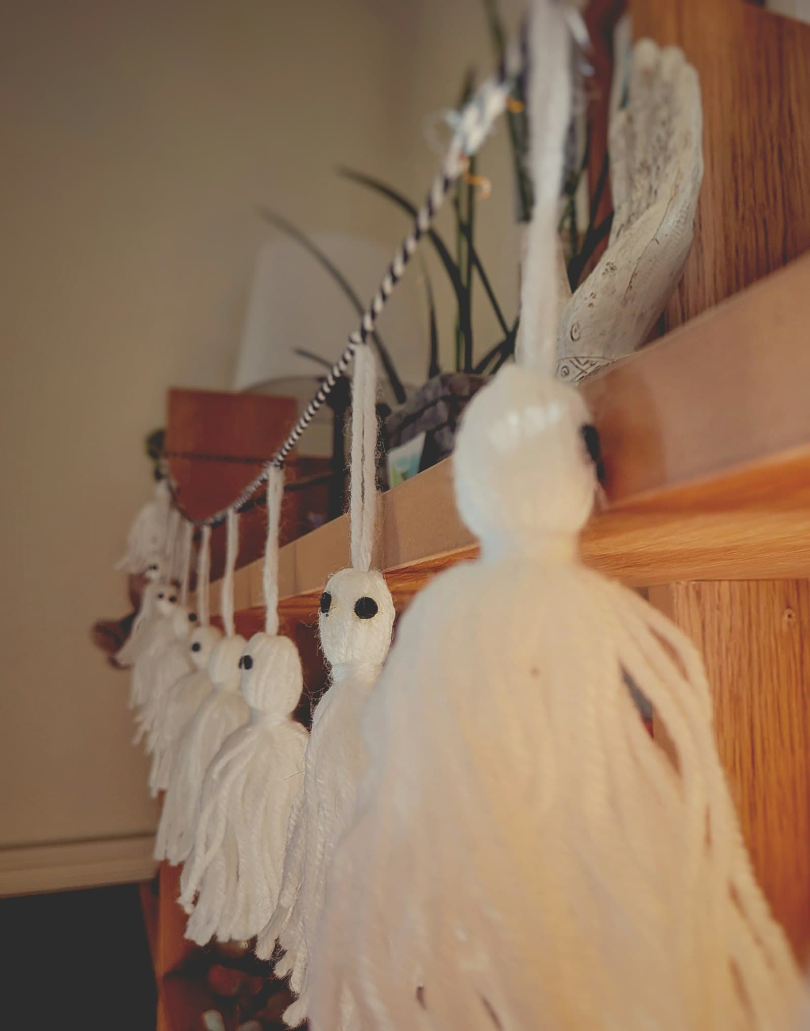 Handmade white yarn ghost decorations with button eyes, hanging in a row from a wooden shelf
